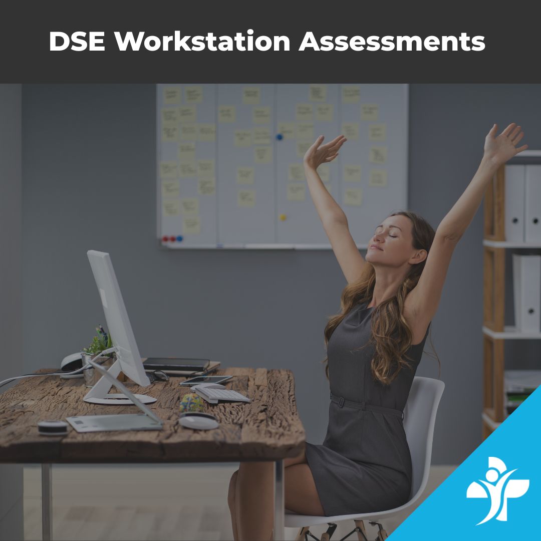 Woman in a gray dress sits at a wooden desk with a computer, stretching her arms overhead during a DSE workstation assessment in a modern office. A memo board with sticky notes is in the background.