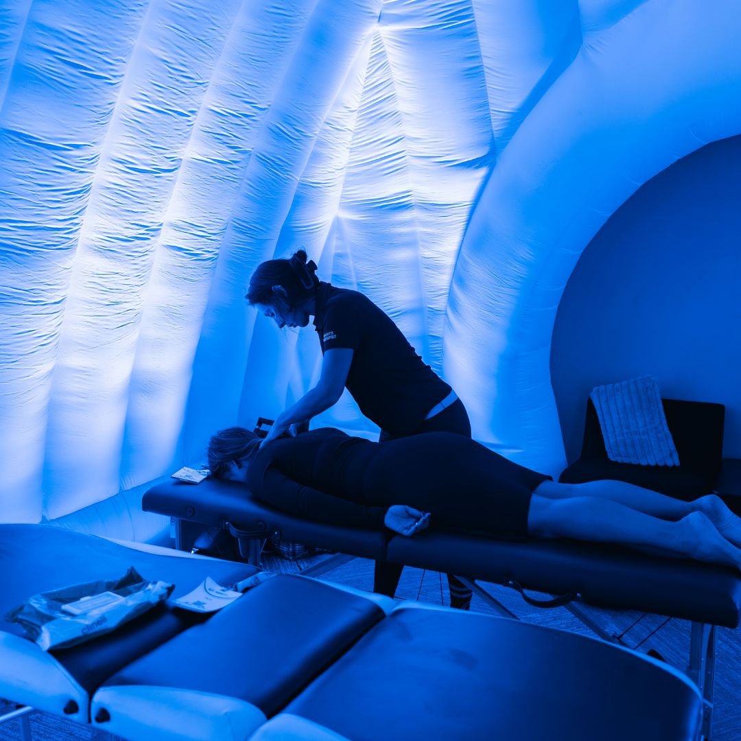 Therapist giving a back massage to a person lying face down on a portable table inside a blue-lit inflatable tent