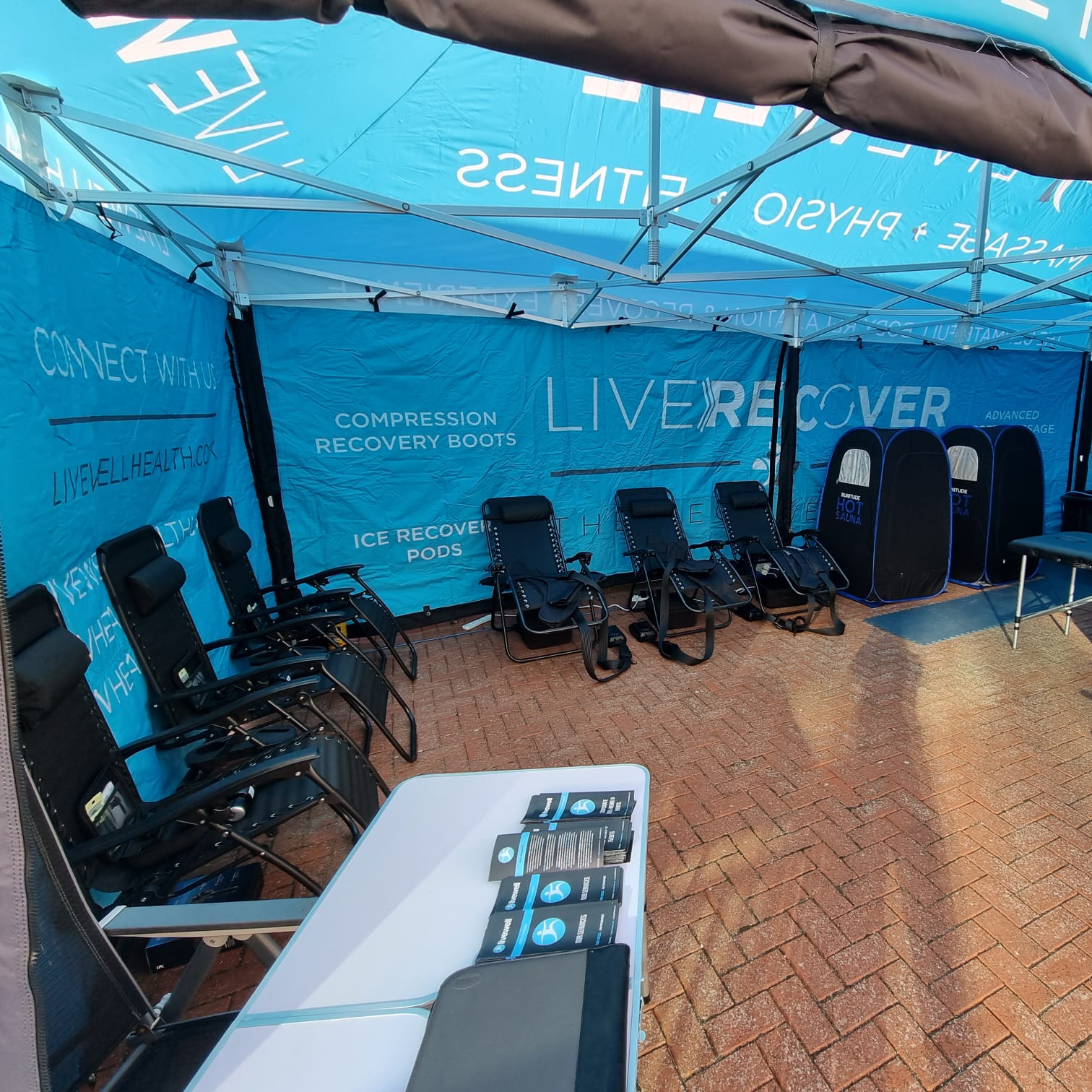 Blue exhibition tent promoting compression recovery boots, with a row of black reclining chairs and a table with brochures on a brick-paved floor