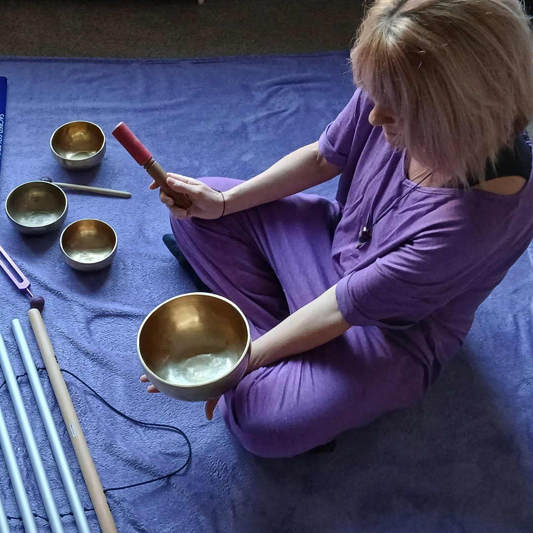Woman in a purple outfit sits on a blue mat, holding a singing bowl and mallet, with several bowls nearby.