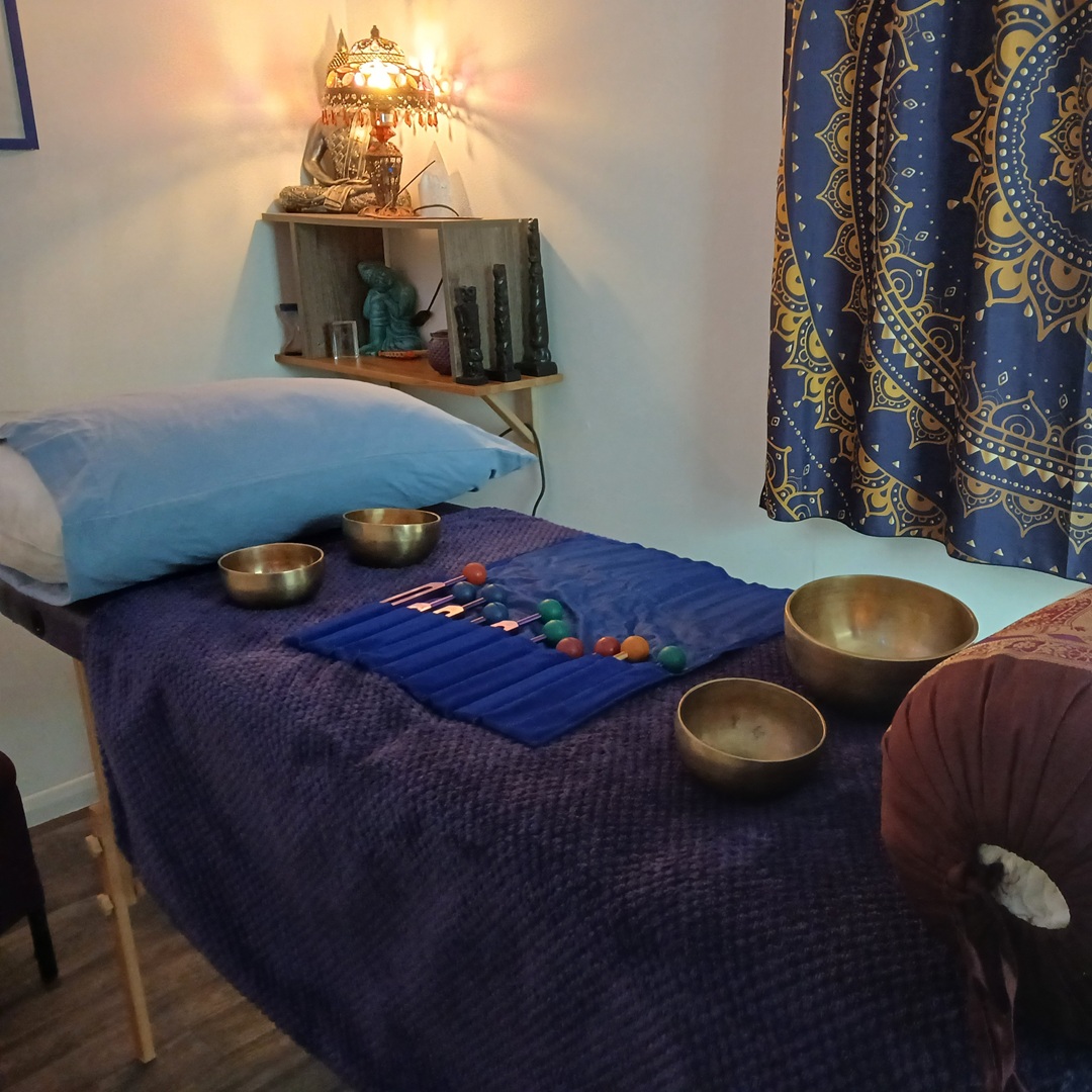 Massage/therapy room setup with singing bowls on a dark blue cloth-covered table and a blue pillow nearby, warm lamp on a shelf in the background.