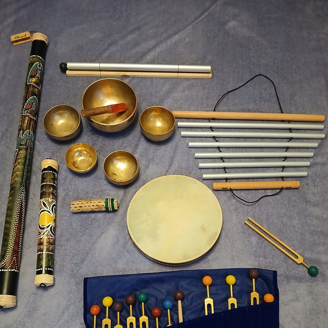 Assorted percussion instruments arranged on a blue carpet: decorated didgeridoo, brass singing bowls, mallets, tuning forks, a frame drum, and a blue roll with colorful mallet heads.