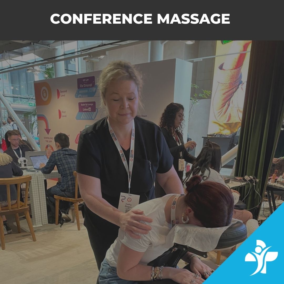 Professional masseuse giving a seated shoulder massage to a conference attendee at an expo, both wearing name badges in a busy venue with booths nearby.