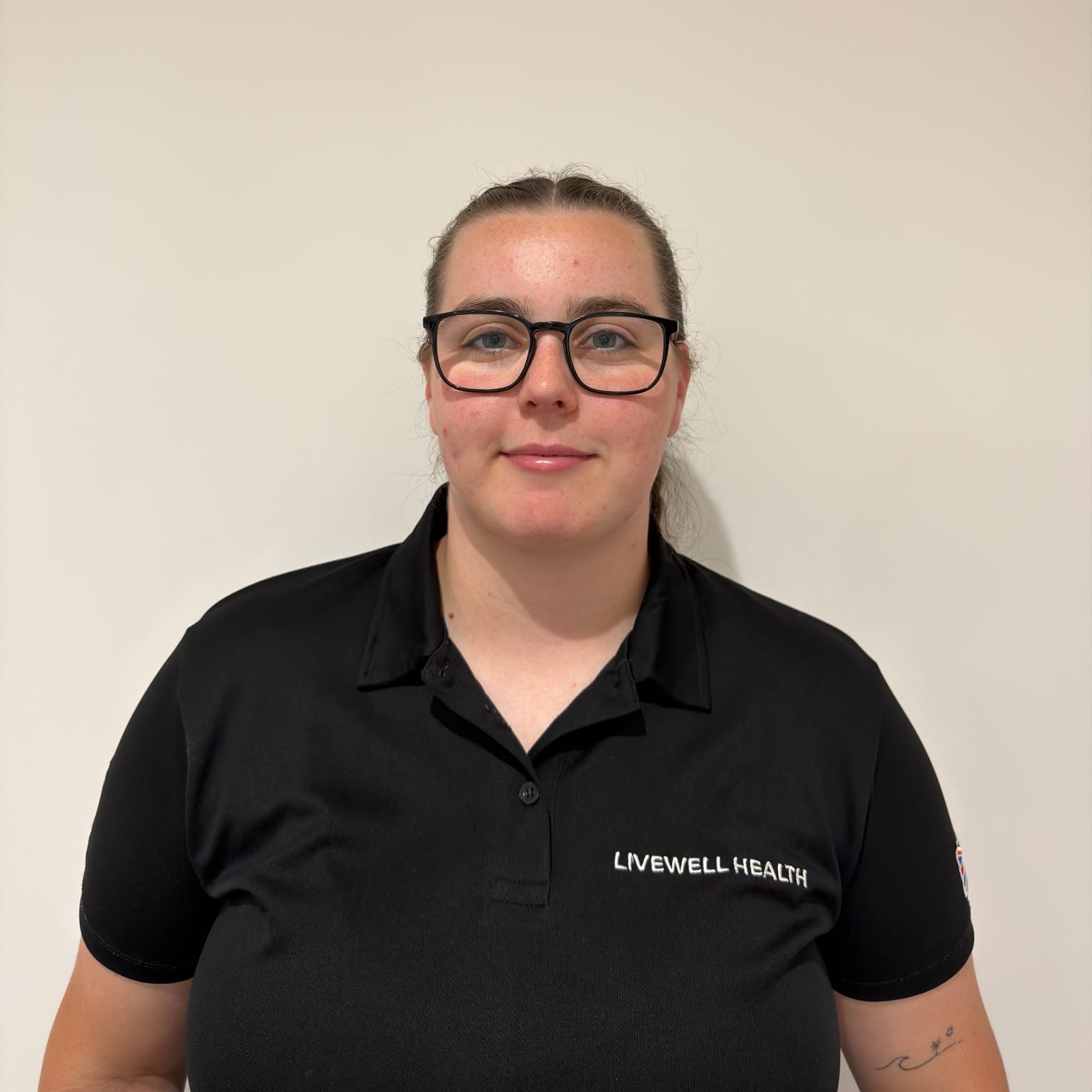 Front-facing woman wearing black polo with Livewell Health logo, glasses, and a ponytail, standing against a light wall.