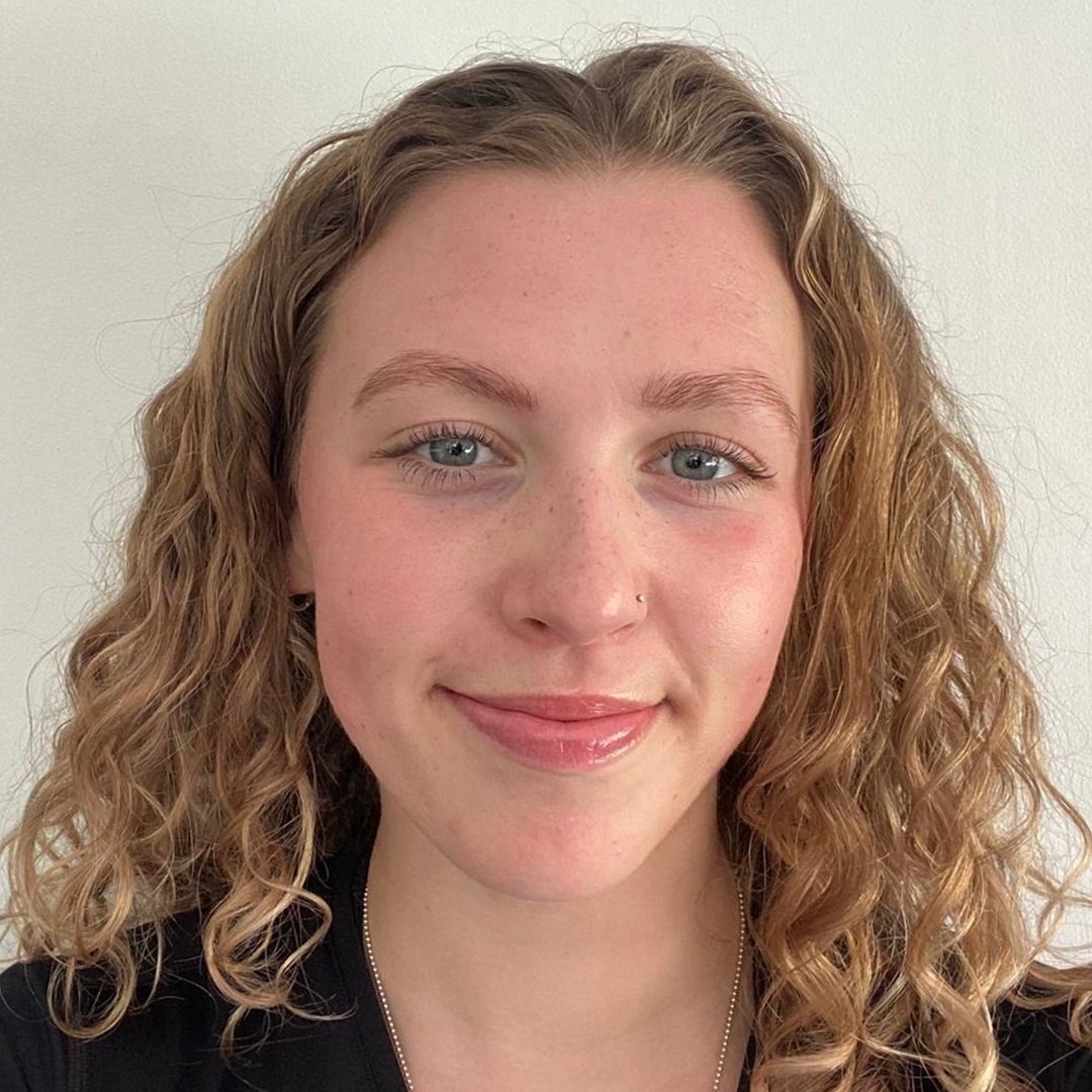 Close-up portrait of a smiling young woman with curly blonde hair, blue eyes, and a nose stud against a light background.